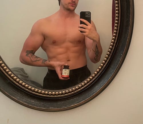 Customer's muscular arm holding an Ancestral Testosterone Booster bottle while posing in a mirror.