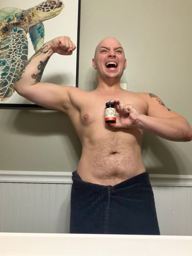 Reviewer flexing a bicep while holding a bottle of Primal Reindeer Testosterone Support in a bathroom.