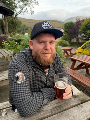 Reviewer in a plaid shirt and baseball cap holding a glass of dark liquid outdoors, reviewing Primal Testosterone Booster.